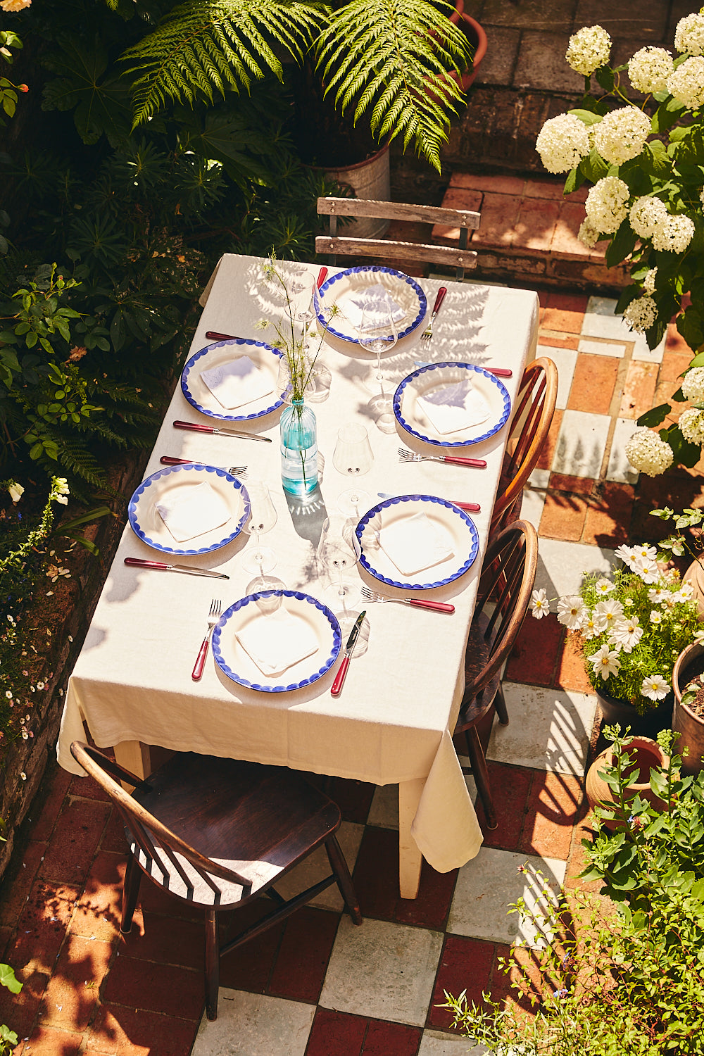 Dining table set for a meal outdoors with plates, cutlery, and glasses on a checkered patio.