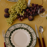 Dinner setting with a decorative plate, grapes, figs, and cutlery on a yellow tablecloth.