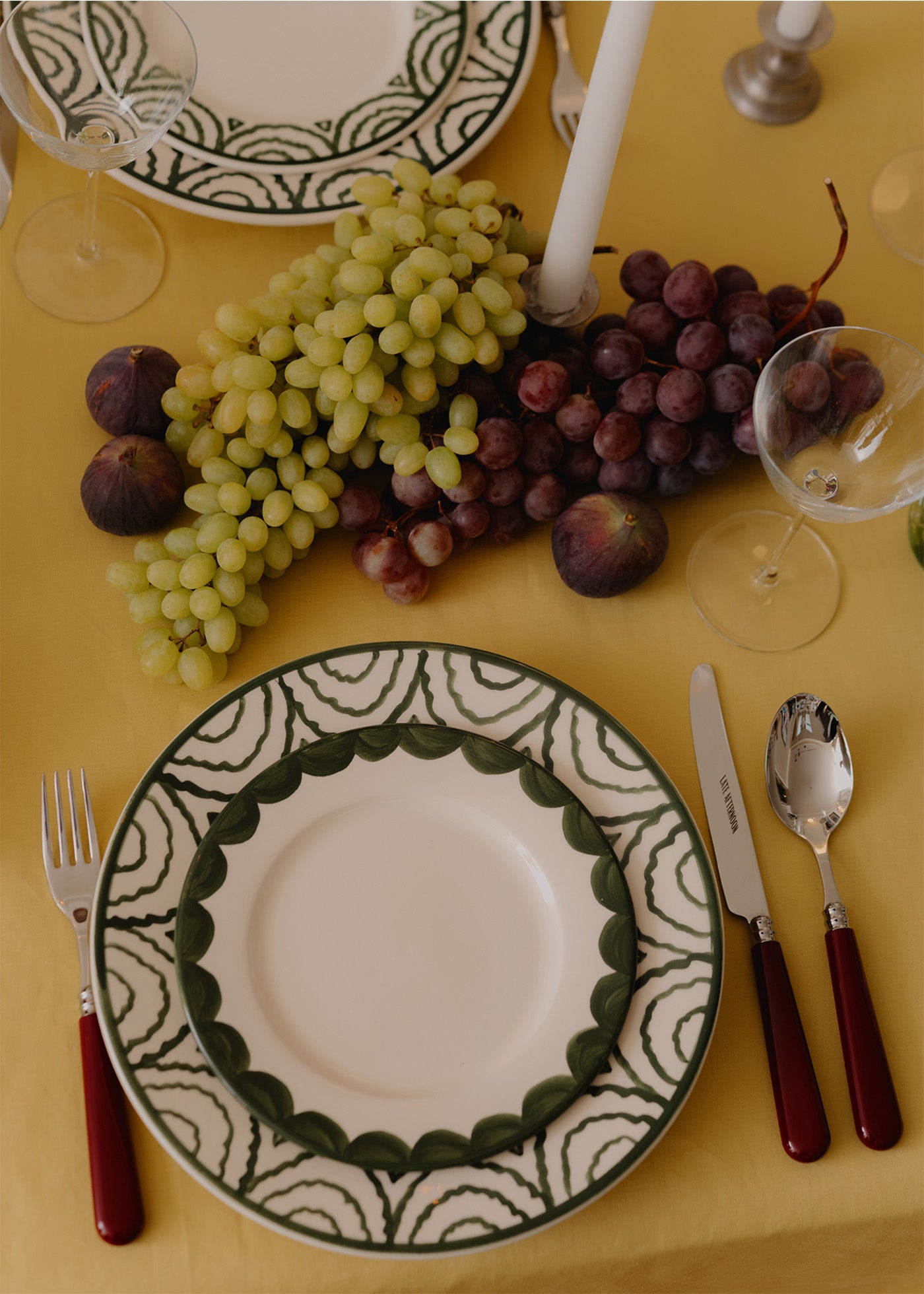 Dinner setting with a decorative plate, grapes, figs, and cutlery on a yellow tablecloth.