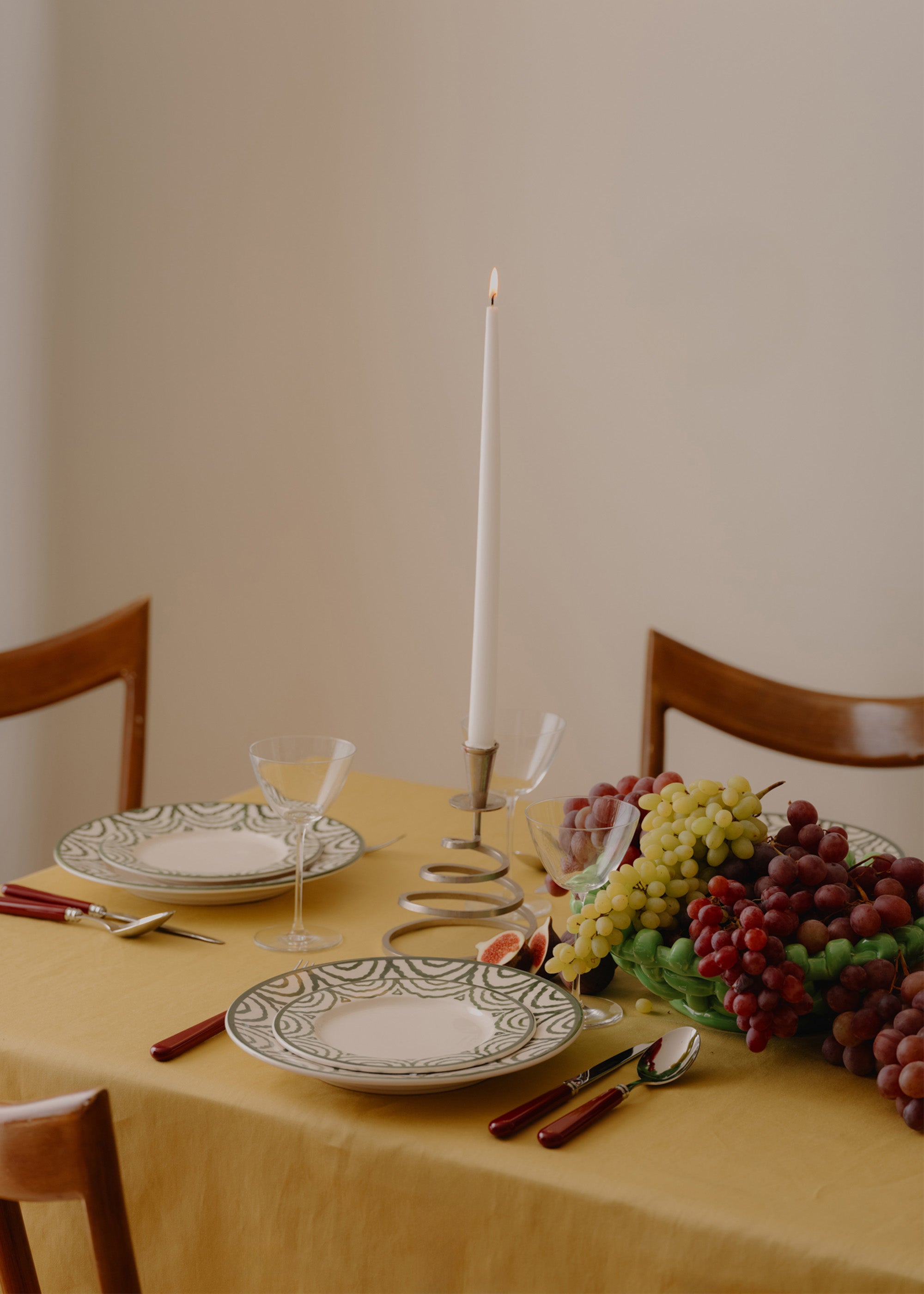 Dining table set with plates, glasses, candles, and grapes on a yellow tablecloth.