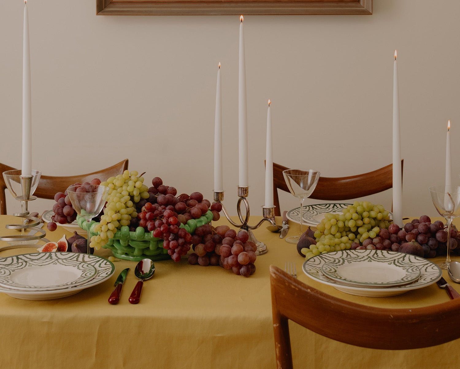 Dining table set with grapes, candles, and plates under a framed painting on the wall.