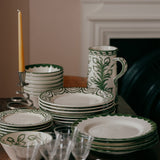 Dining table set with green and white patterned plates, cups, and glasses in front of a fireplace.