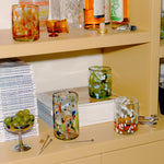 A styled shelf display featuring several Late afternoon colorful hand-blown glass tumblers with speckled patterns, alongside bottles, books, a bowl of green olives, and cocktail tools in a warm beige interior setting.