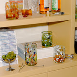 A styled shelf display featuring several Late afternoon colorful hand-blown glass tumblers with speckled patterns, alongside bottles, books, a bowl of green olives, and cocktail tools in a warm beige interior setting.