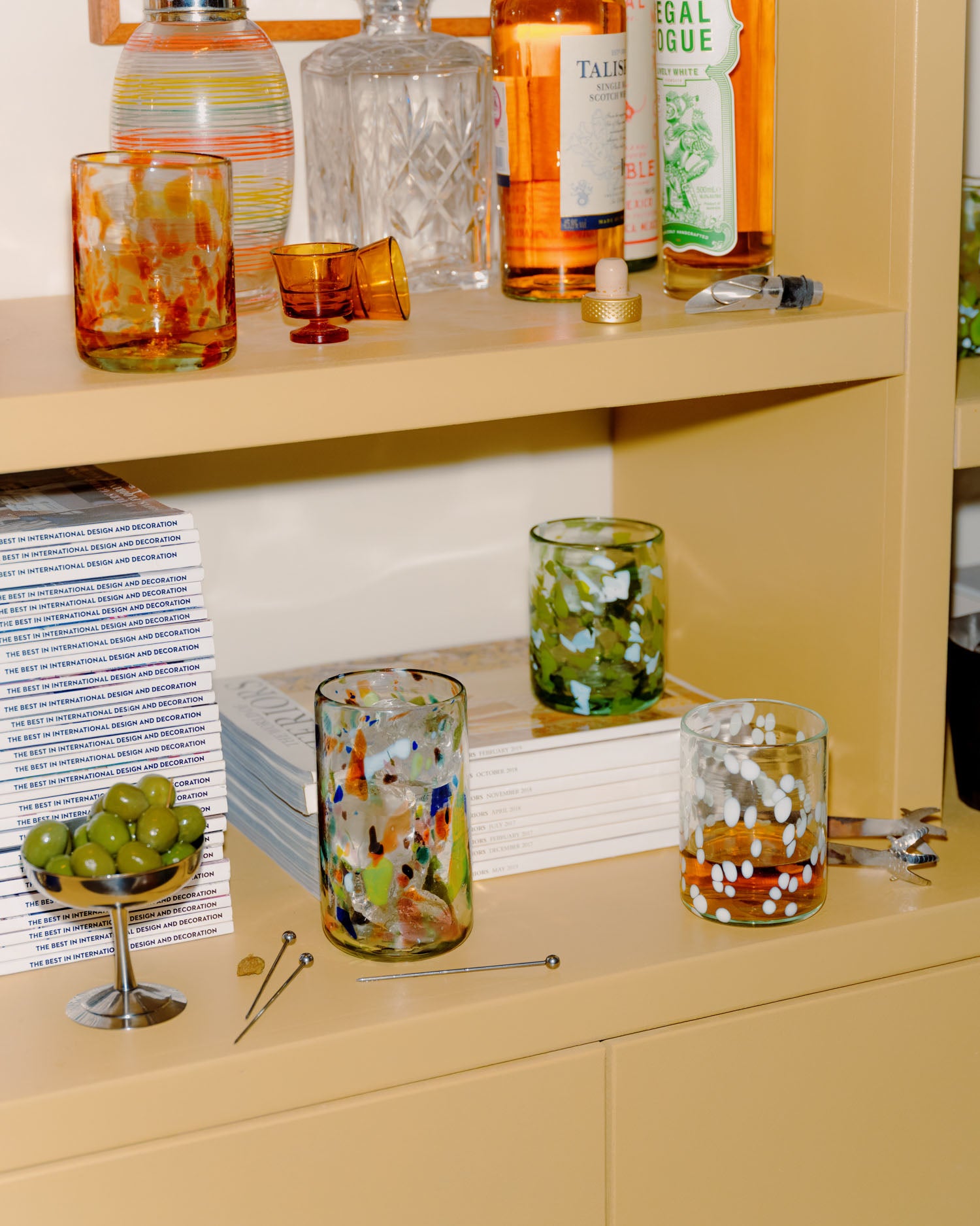 A styled shelf display featuring several Late afternoon colorful hand-blown glass tumblers with speckled patterns, alongside bottles, books, a bowl of green olives, and cocktail tools in a warm beige interior setting.