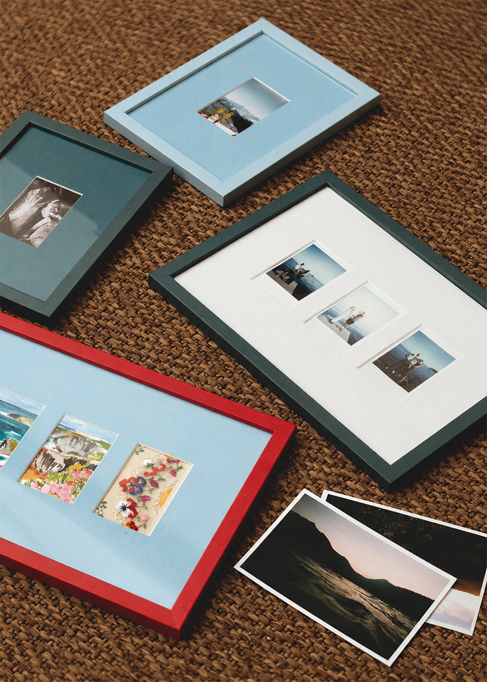 Four picture frames in red, dark teal, and light blue, arranged on a brown woven rug with sample photographs inside and beside them.