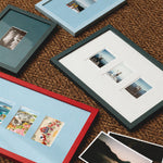 Four picture frames in red, dark teal, and light blue, arranged on a brown woven rug with sample photographs inside and beside them.