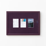 A square dark purple framed art piece mounted on a white wall, featuring three small vertical printed inserts with handwritten notes and a landscape photograph.
