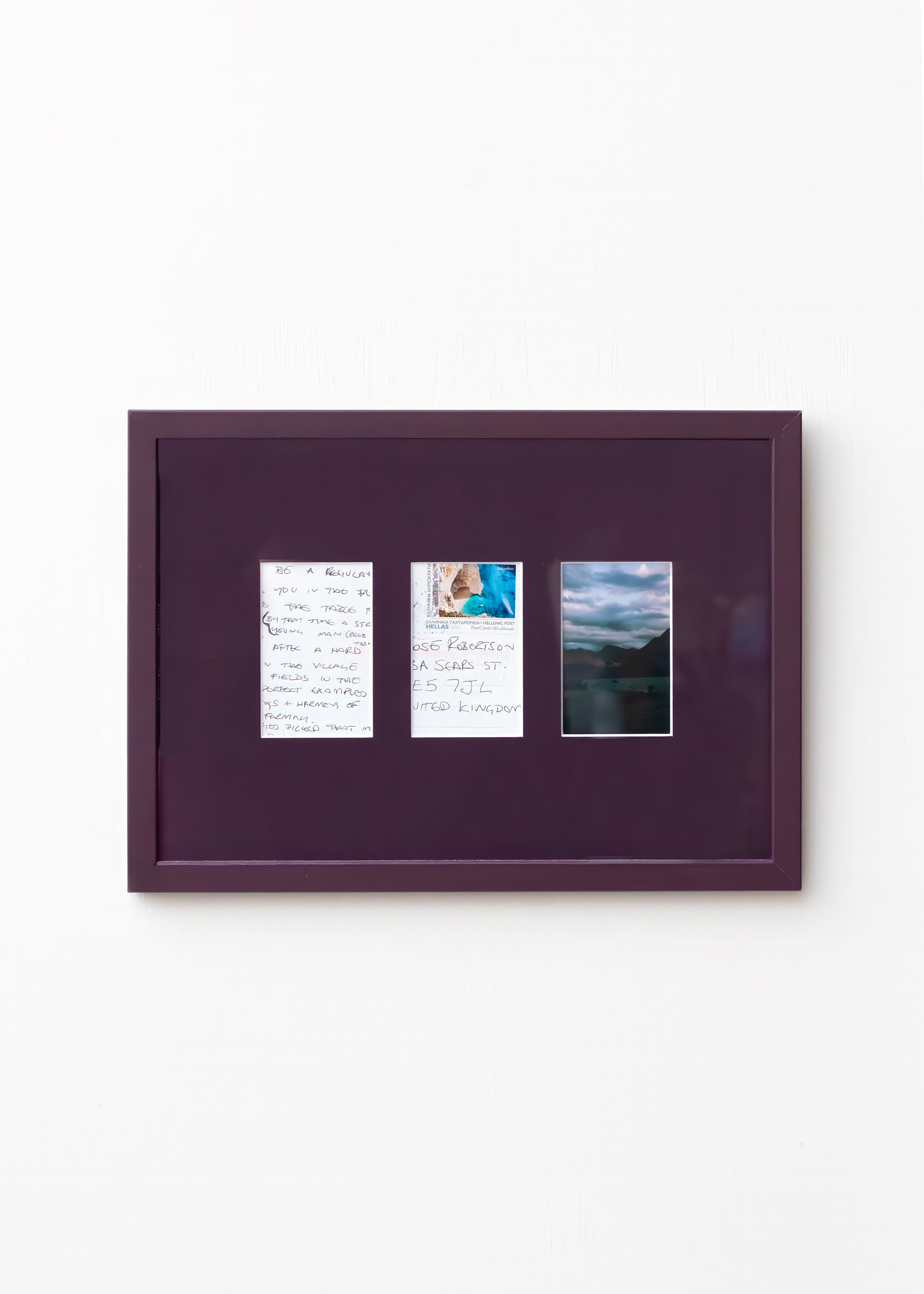 A square dark purple framed art piece mounted on a white wall, featuring three small vertical printed inserts with handwritten notes and a landscape photograph.