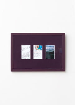 A square dark purple framed art piece mounted on a white wall, featuring three small vertical printed inserts with handwritten notes and a landscape photograph.