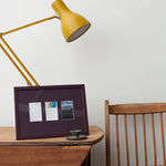 A dark purple picture frame with three small printed cards or photos inside, styled on a wooden table next to a yellow desk lamp and a wooden chair against a white wall.