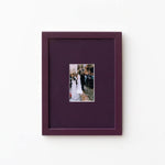 A square dark purple picture frame with a matching mat, featuring a small central photograph of a bride and groom, displayed on a clean white wall.