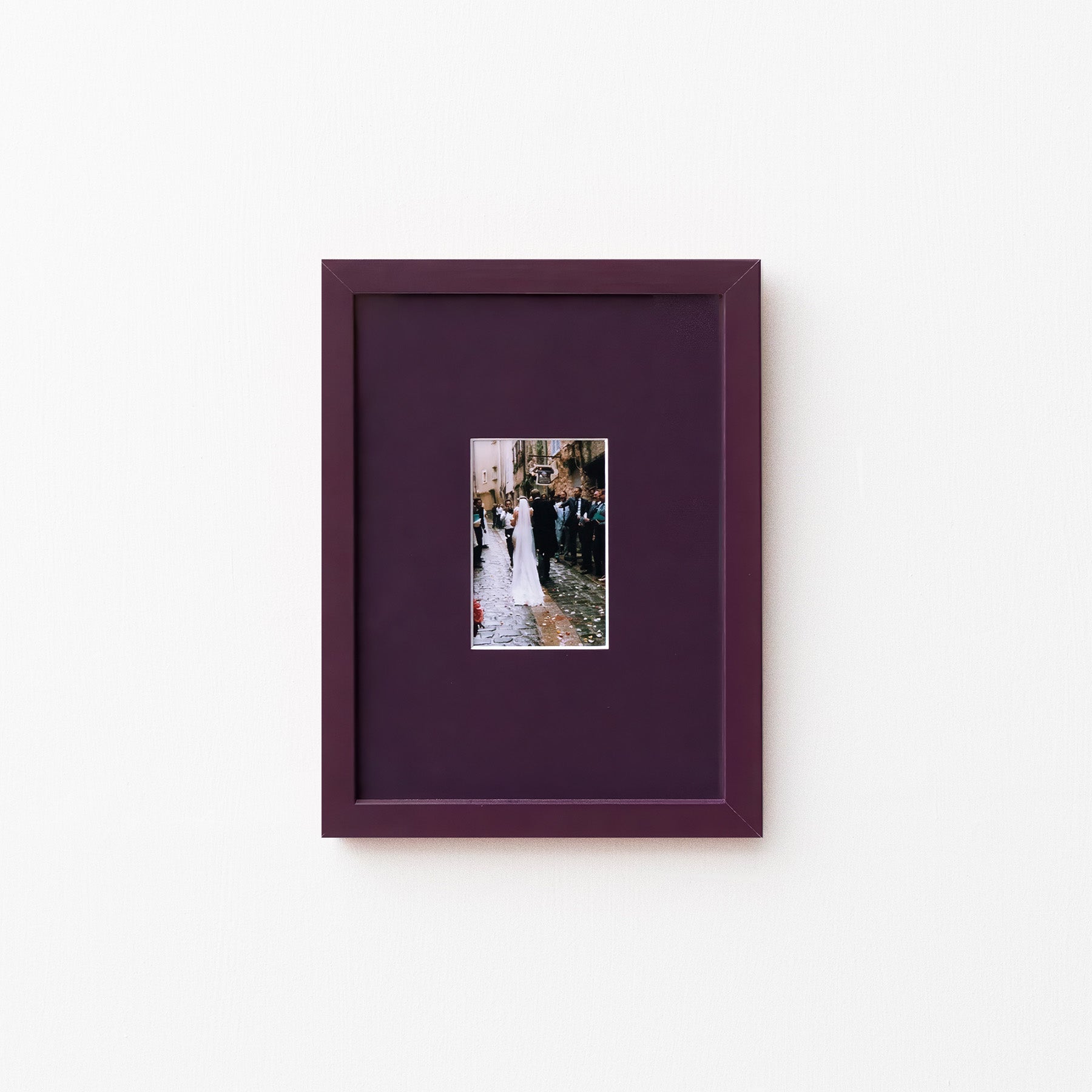 A square dark purple picture frame with a matching mat, featuring a small central photograph of a bride and groom, displayed on a clean white wall.