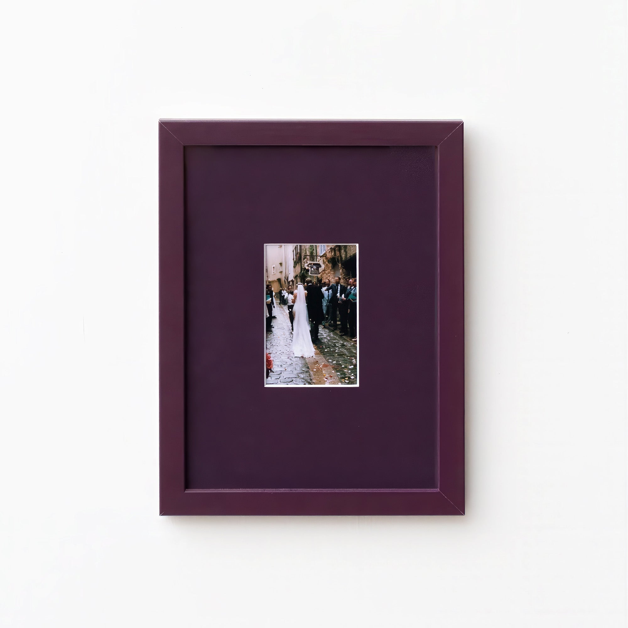A square dark purple picture frame with a matching mat, featuring a small central photograph of a bride and groom, displayed on a clean white wall.