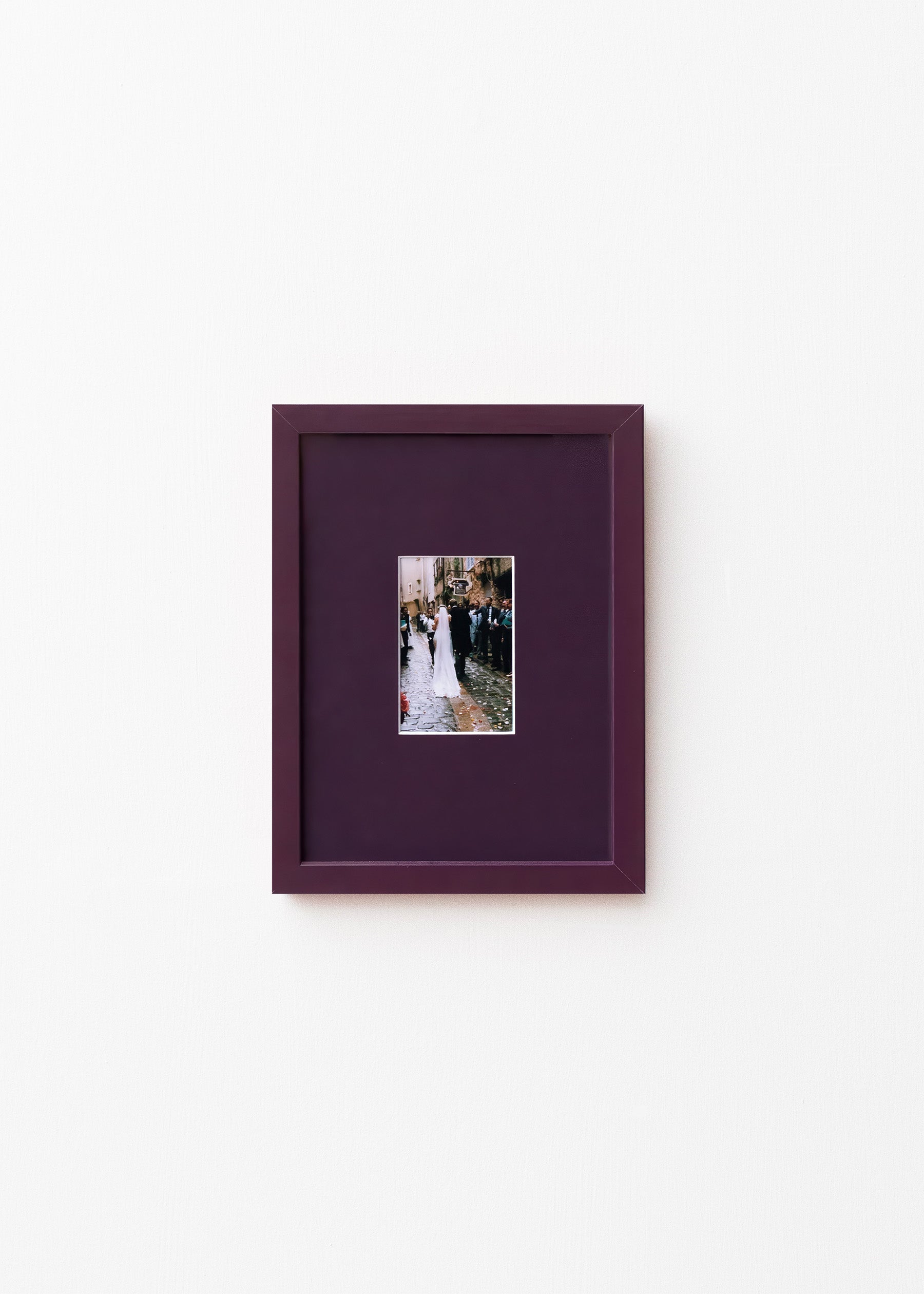 A square dark purple picture frame with a matching mat, featuring a small central photograph of a bride and groom, displayed on a clean white wall.