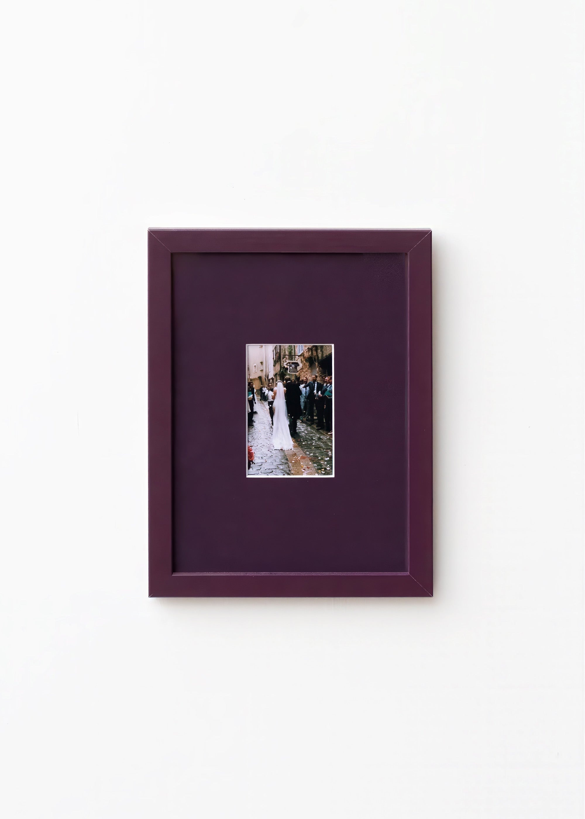 A square dark purple picture frame with a matching mat, featuring a small central photograph of a bride and groom, displayed on a clean white wall.