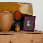 A styled interior scene showing a wooden dresser with a large terracotta vase, a small ceramic jar, a framed photo, and an abstract painting leaning against a white wall.