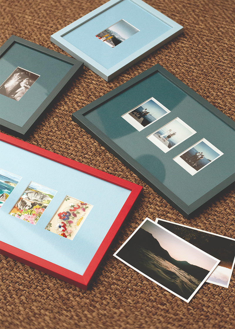 Four picture frames in different colors and sizes arranged on a brown woven rug, with several printed photographs placed beside them.