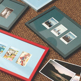 Four picture frames in different colors and sizes arranged on a brown woven rug, with several printed photographs placed beside them.