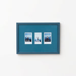 A rectangular Petrol Blue picture frame with three small printed photographs mounted side by side, shown hanging on a plain white wall.