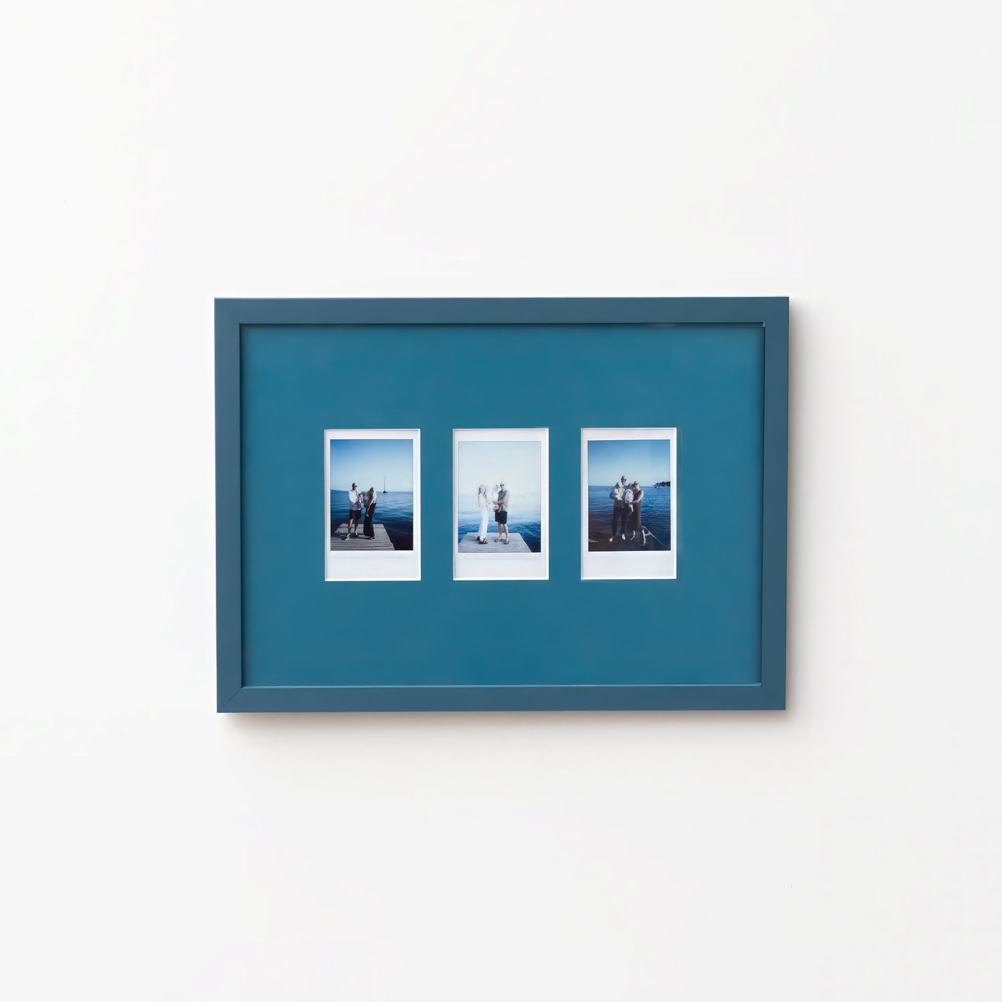 A rectangular Petrol Blue picture frame with three small printed photographs mounted side by side, shown hanging on a plain white wall.