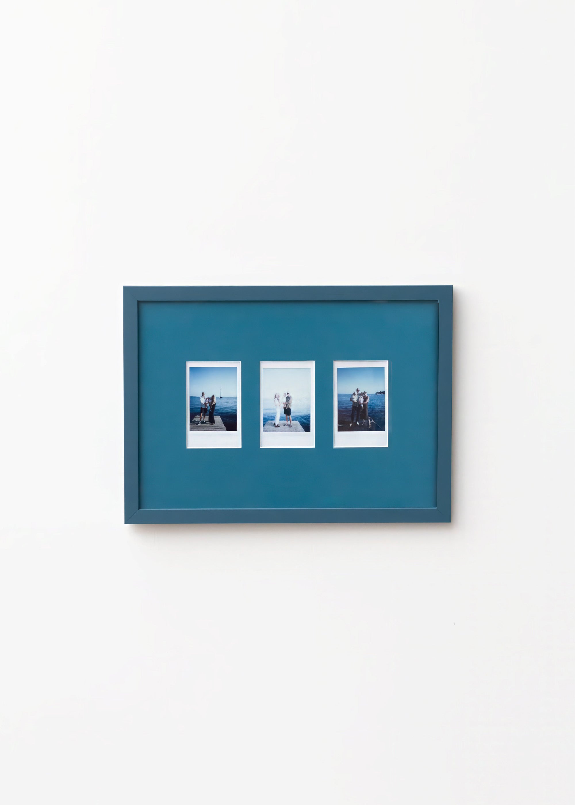 A rectangular Petrol Blue picture frame with three small printed photographs mounted side by side, shown hanging on a plain white wall.