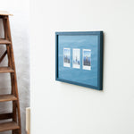 A framed wall art piece with three small vertical landscape photographs mounted on a blue textured background, hung on a white wall next to a wooden ladder.