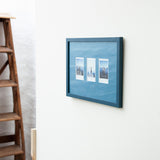A framed wall art piece with three small vertical landscape photographs mounted on a blue textured background, hung on a white wall next to a wooden ladder.