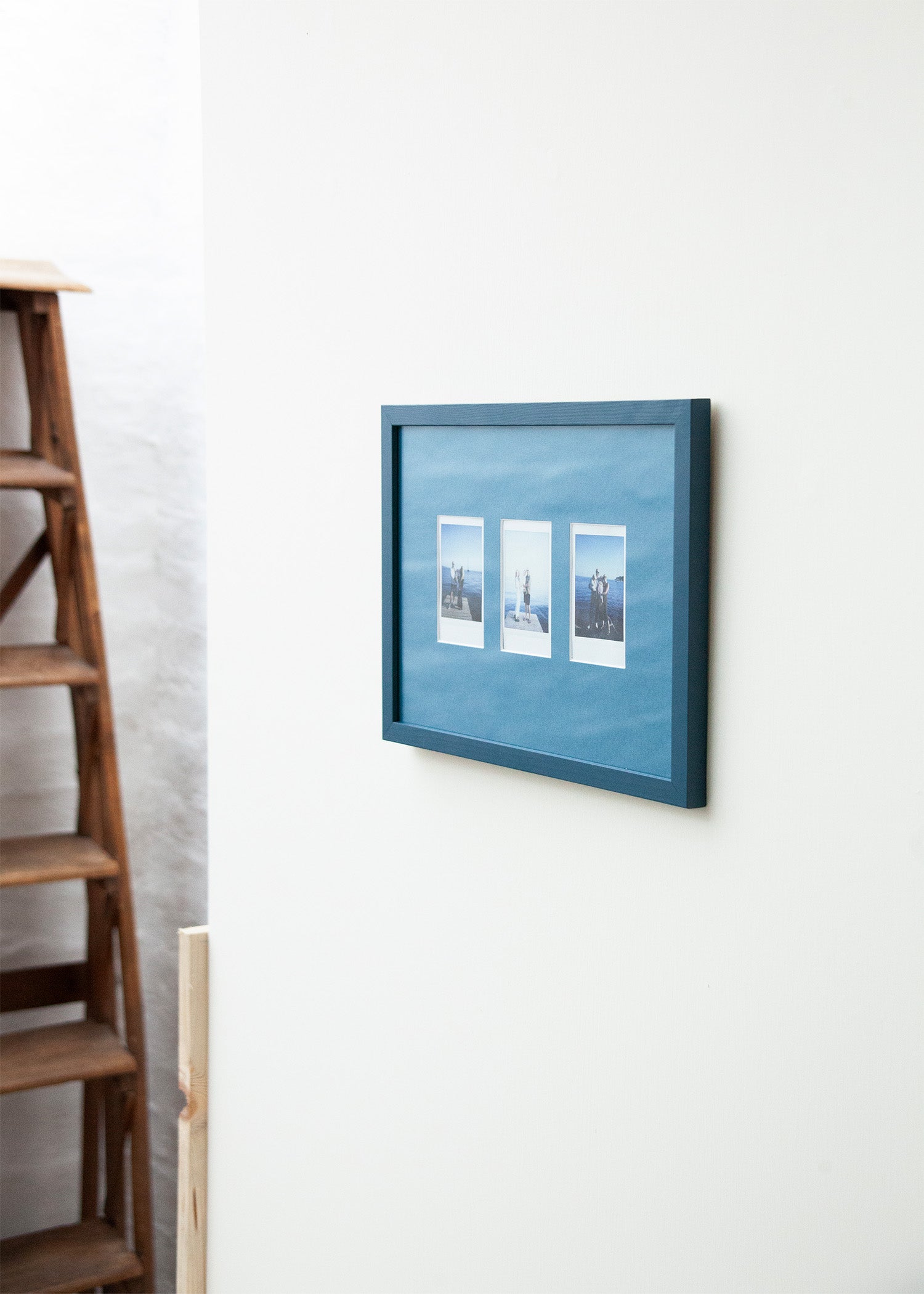A framed wall art piece with three small vertical landscape photographs mounted on a blue textured background, hung on a white wall next to a wooden ladder.