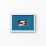 A square framed art print featuring a black and white ultrasound photo of a baby, set against a teal-blue background with a light blue frame, mounted on a plain white wall.