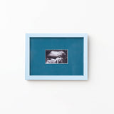 A square framed art print featuring a black and white ultrasound photo of a baby, set against a teal-blue background with a light blue frame, mounted on a plain white wall.