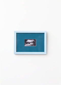 A square framed art print featuring a black and white ultrasound photo of a baby, set against a teal-blue background with a light blue frame, mounted on a plain white wall.