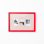 A picture frame with a bright red border and a pale pink mat, holding three small vertical photographs, shown against a plain white wall.