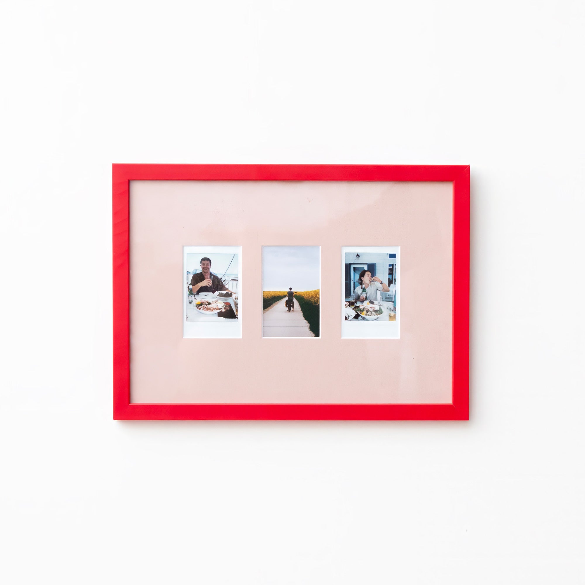 A picture frame with a bright red border and a pale pink mat, holding three small vertical photographs, shown against a plain white wall.