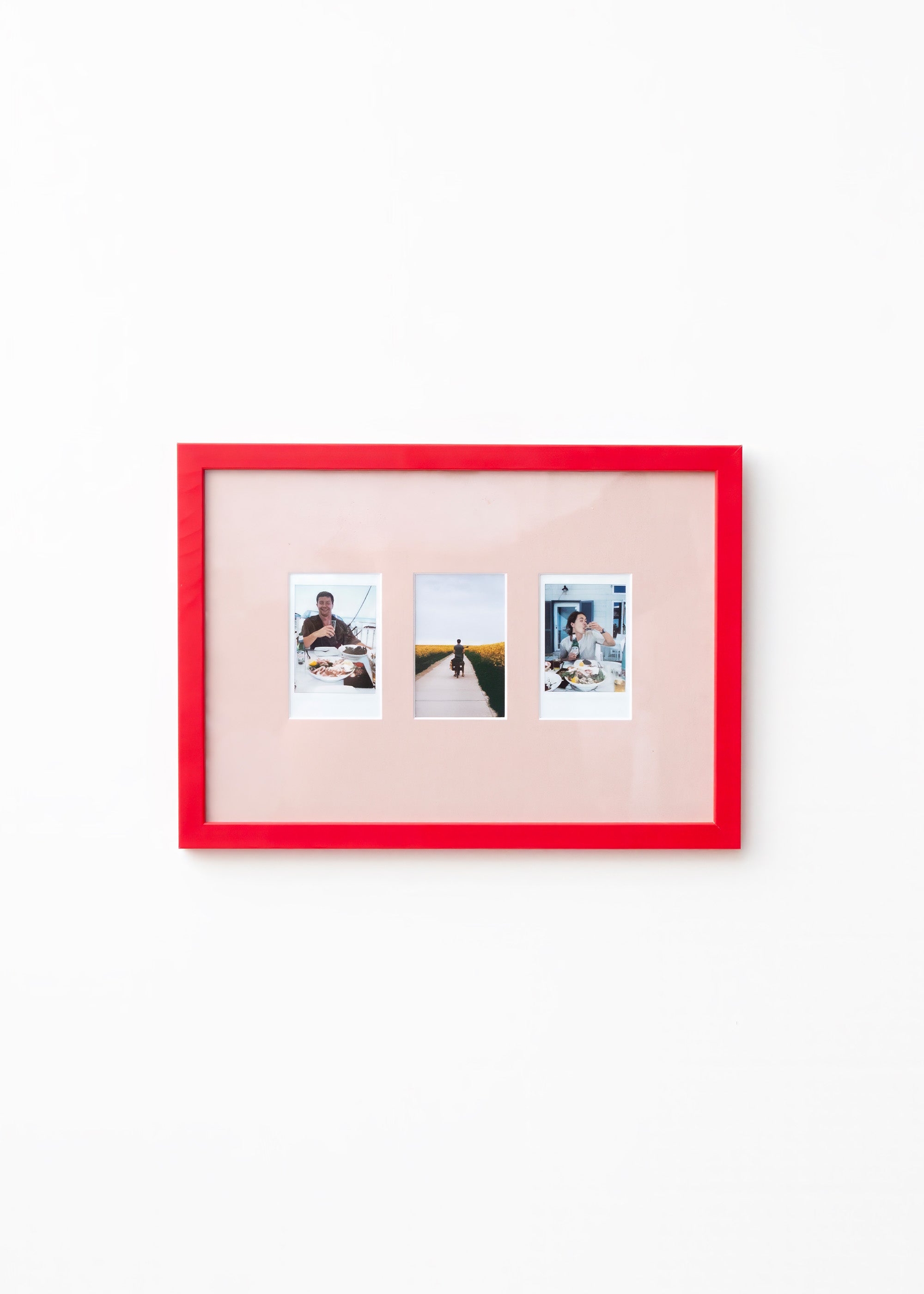 A picture frame with a bright red border and a pale pink mat, holding three small vertical photographs, shown against a plain white wall.
