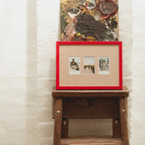 A framed art print with a bright red frame and beige matting, containing three small photographs, displayed on a rustic wooden stool against a white textured wall. Above the frame sits a textured abstract painting.