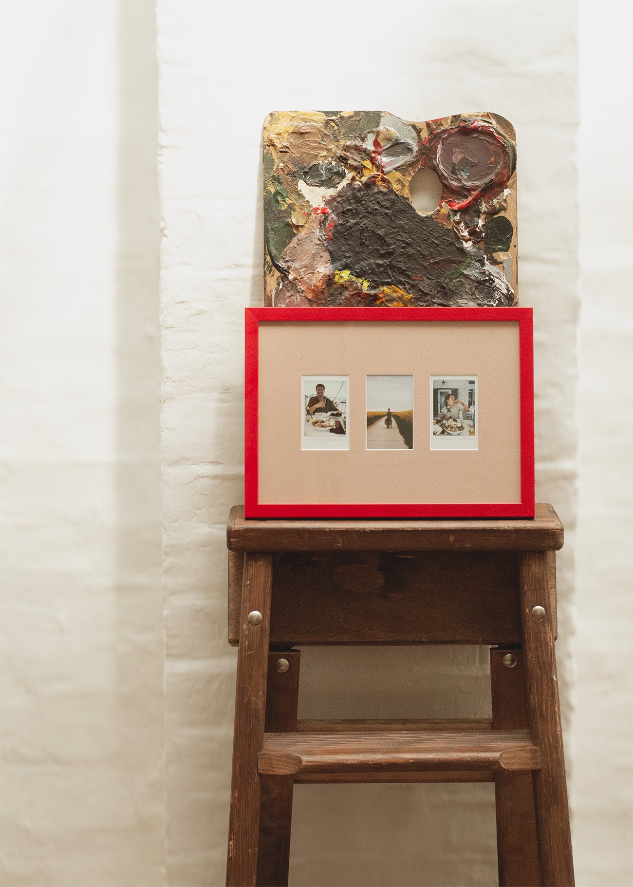 A framed art print with a bright red frame and beige matting, containing three small photographs, displayed on a rustic wooden stool against a white textured wall. Above the frame sits a textured abstract painting.