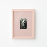 A vertical light pink picture frame with a white mat, showcasing a small black and white portrait photograph, mounted on a clean white wall.
