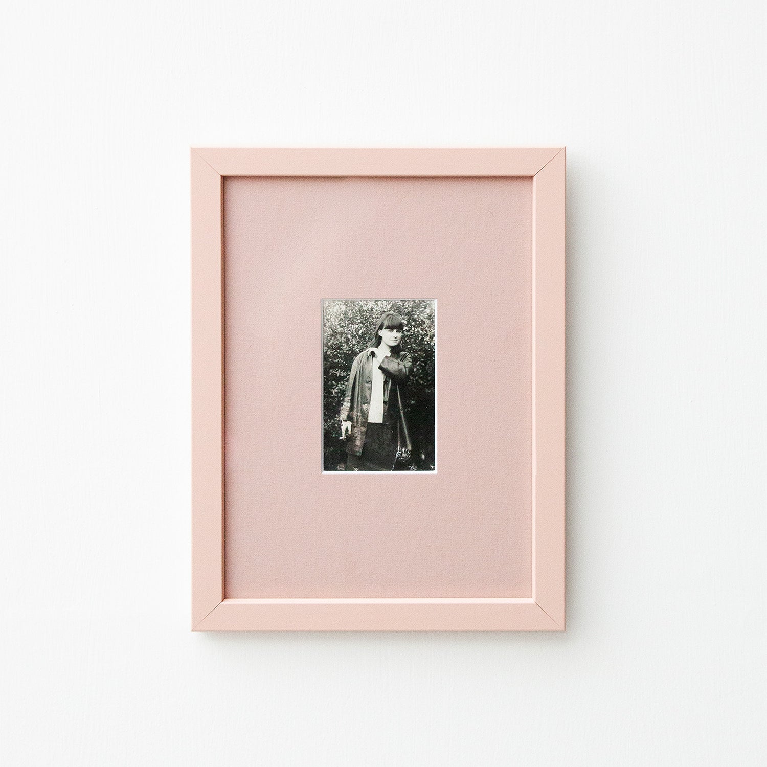 A vertical light pink picture frame with a white mat, showcasing a small black and white portrait photograph, mounted on a clean white wall.