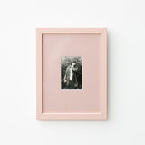A vertical light pink picture frame with a white mat, showcasing a small black and white portrait photograph, mounted on a clean white wall.