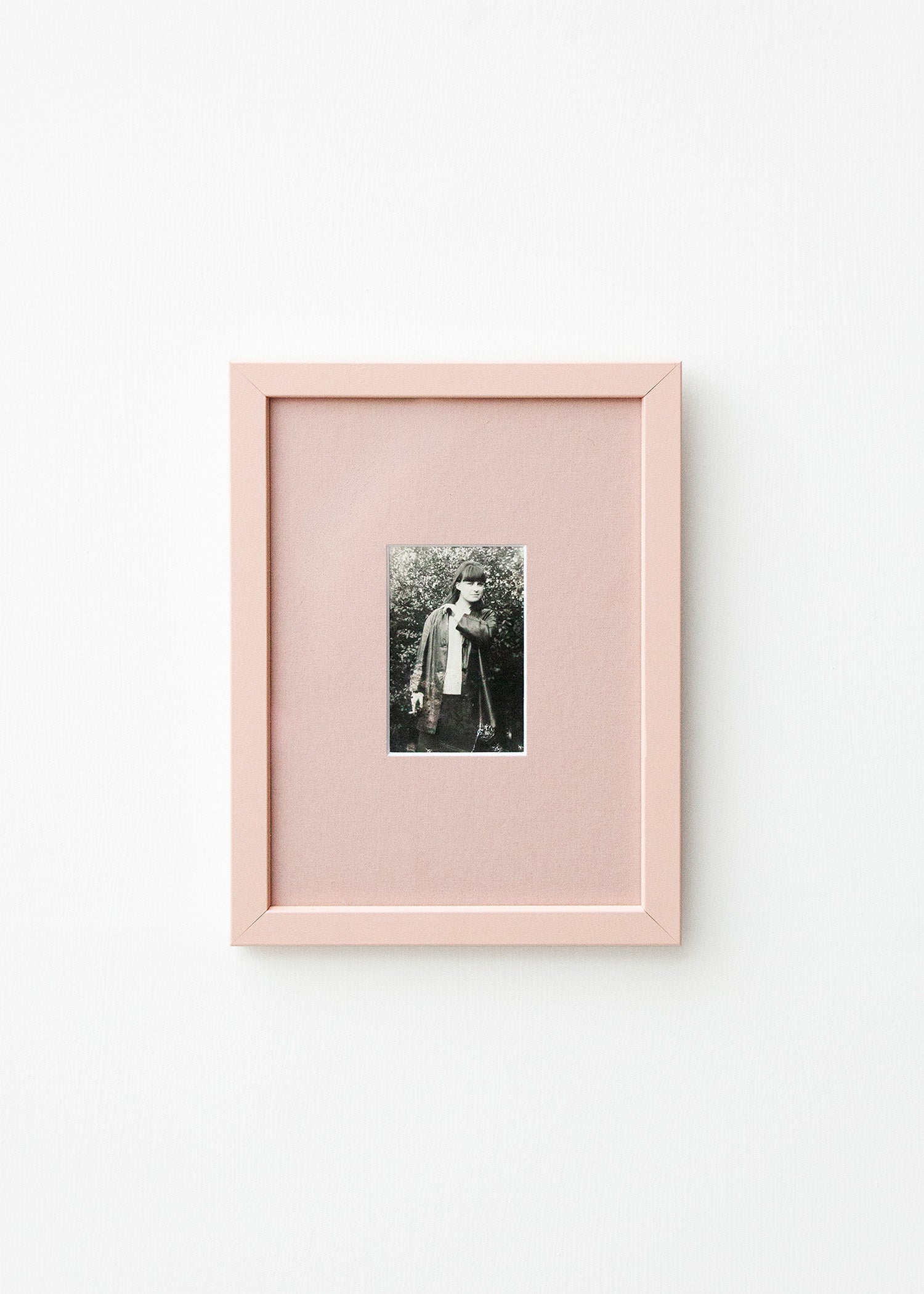 A vertical light pink picture frame with a white mat, showcasing a small black and white portrait photograph, mounted on a clean white wall.
