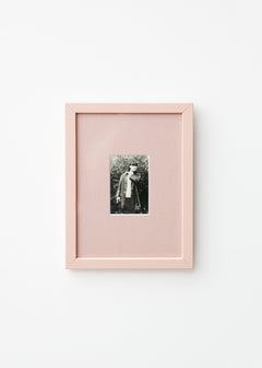 A vertical light pink picture frame with a white mat, showcasing a small black and white portrait photograph, mounted on a clean white wall.
