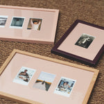 Three rectangular picture frames with pastel pink mats are arranged on a brown woven rug. One frame is light wood, one is dark wood with a deep purple border, and one is pale pink.