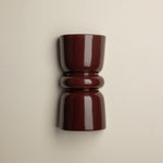 A glossy dark burgundy wall-mounted cup or candle holder with a flared top and ribbed middle section, shown against a plain warm off-white wall.