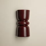 A glossy dark burgundy wall-mounted cup or candle holder with a flared top and ribbed middle section, shown against a plain warm off-white wall.