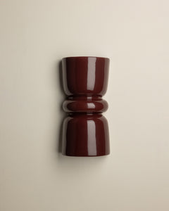 A glossy dark burgundy wall-mounted cup or candle holder with a flared top and ribbed middle section, shown against a plain warm off-white wall.
