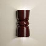 A glossy dark red wall sconce with a sculptural, hourglass-like shape, mounted on a light beige wall and casting a warm glow upwards and downwards.