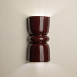 A glossy dark red wall sconce with a sculptural, hourglass-like shape, mounted on a light beige wall and casting a warm glow upwards and downwards.