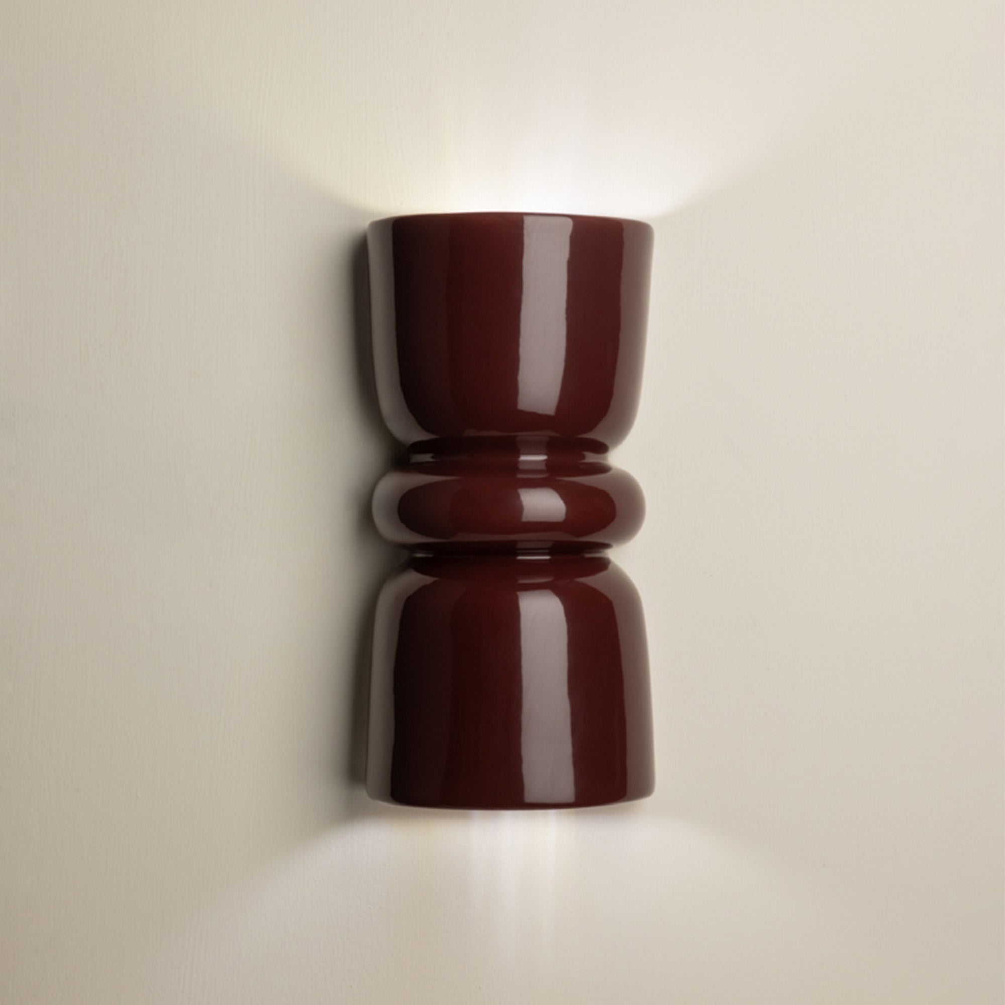 A glossy dark red wall sconce with a sculptural, hourglass-like shape, mounted on a light beige wall and casting a warm glow upwards and downwards.