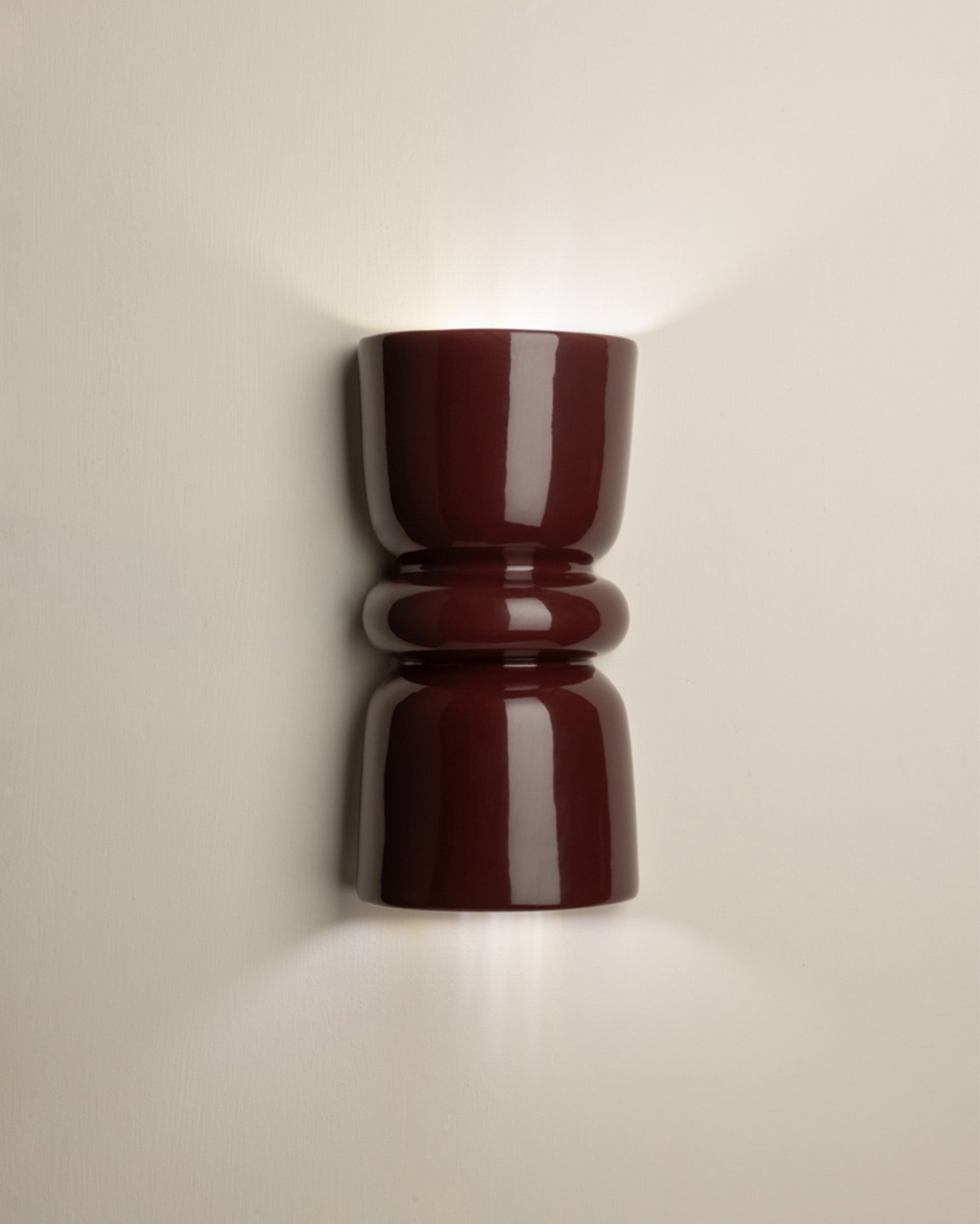A glossy dark red wall sconce with a sculptural, hourglass-like shape, mounted on a light beige wall and casting a warm glow upwards and downwards.
